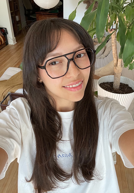 A smiling woman with long dark hair and large black glasses taking a selfie indoors next to a plant.
