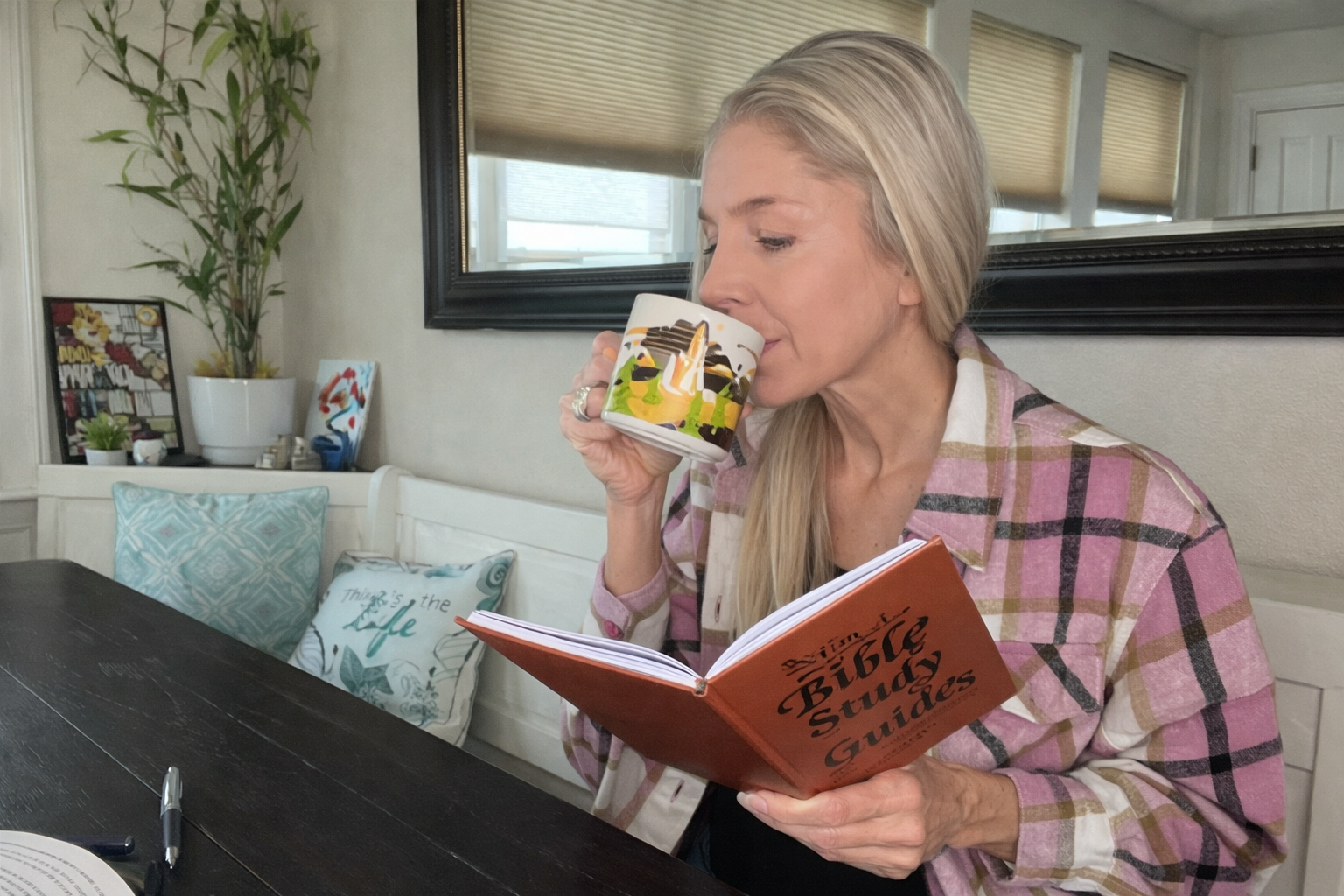 A woman in a plaid shirt sips from a mug while reading a Bible Study guide at a table.