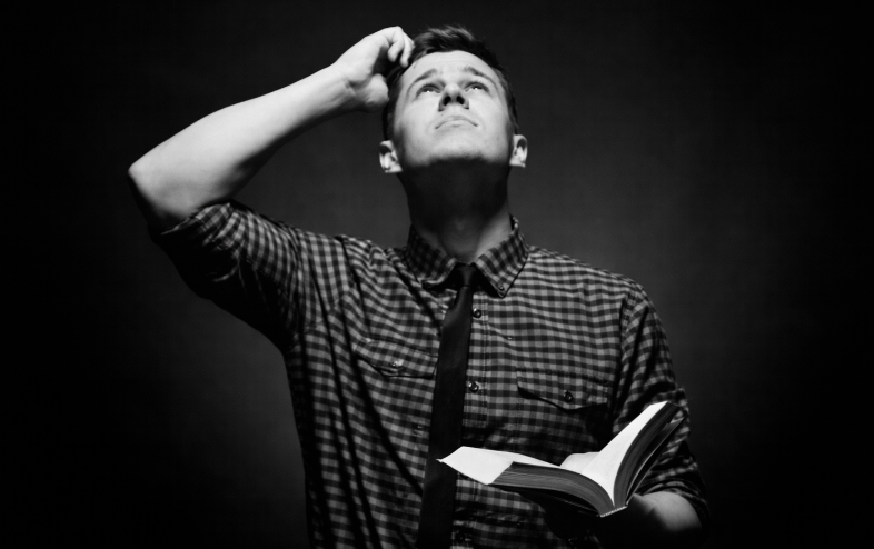 A man in a plaid shirt holds a book, looks up, and scratches his head.