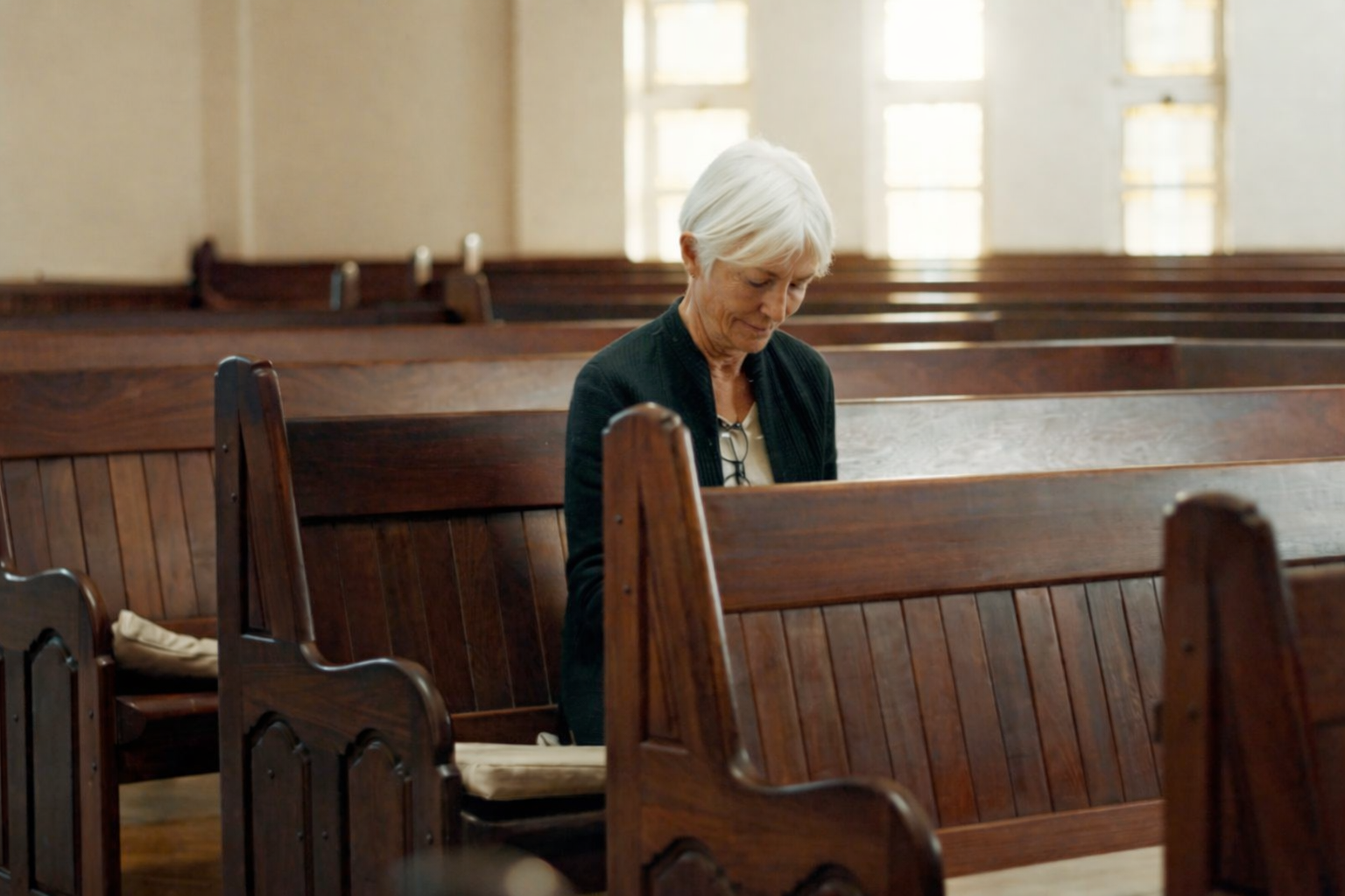An elderly woman with white hair sits alone in a wooden church pew, looking down.
