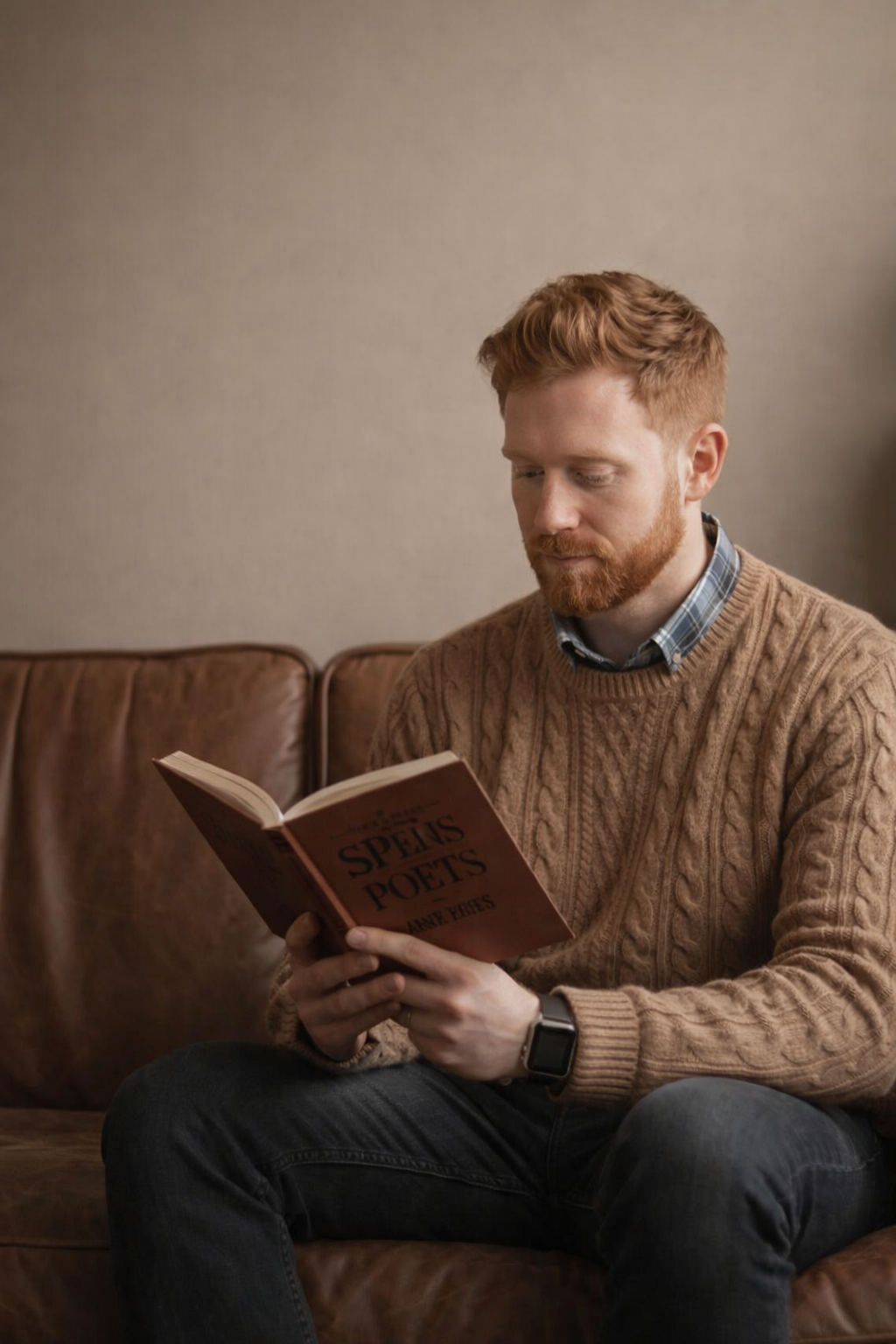 A man with red hair wearing a brown sweater sits on a leather couch reading a book.