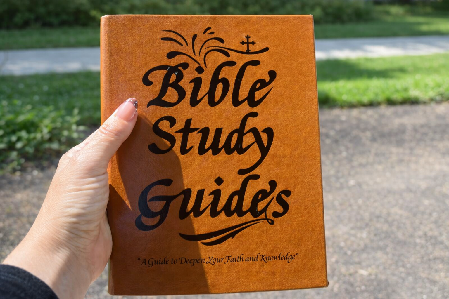 A person's hand holding a tan book titled 'Bible Study Guides' outdoors.