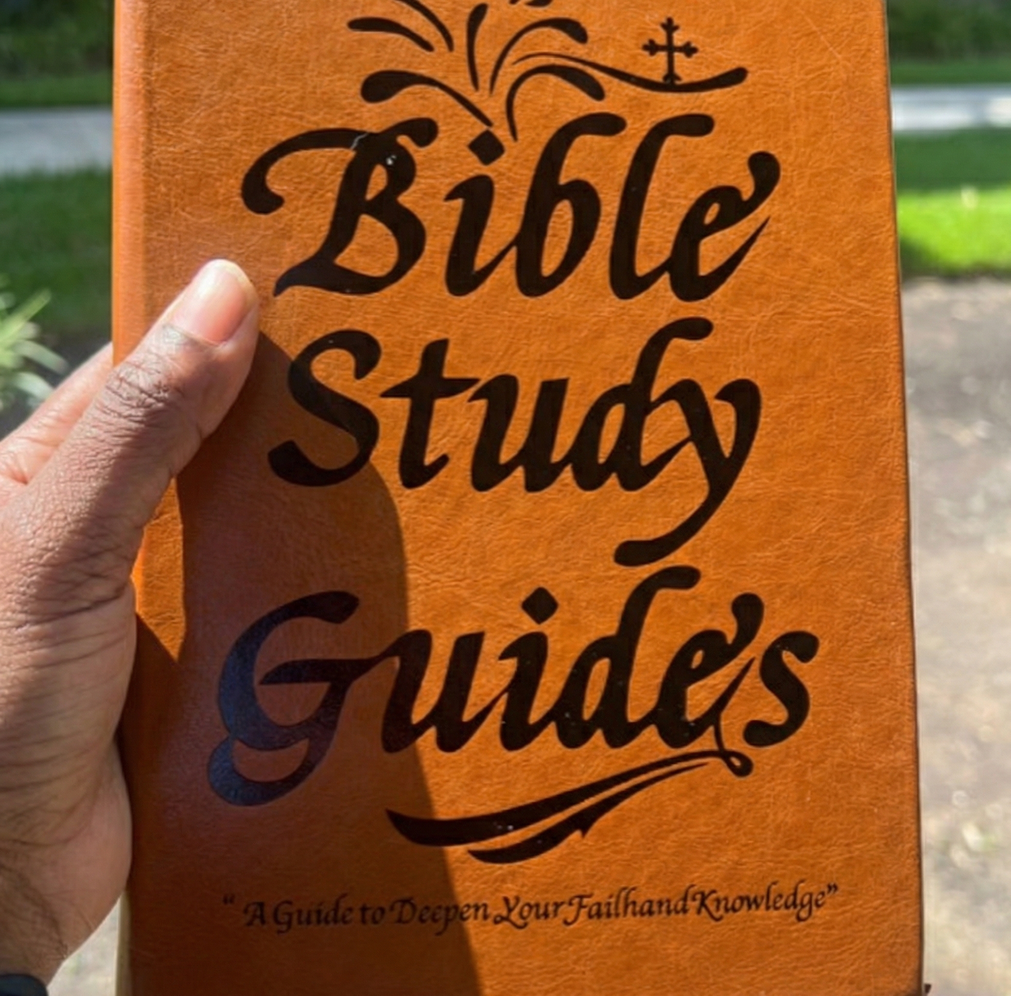 A person's hand holding a brown book titled 'Bible Study Guides' outdoors.
