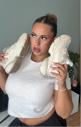 Person holding two sneakers to their ears, wearing a white top.