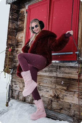 Persoon in winterkleding staand tegen een houten hutwand met sneeuwlaarzen en oorwarmers.