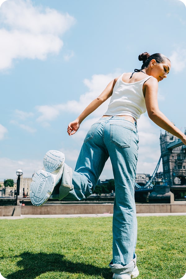 Person in jeans and white top jumping on grass near a bridge.