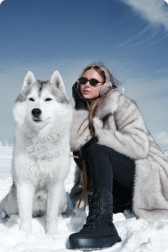 Persoon in bontjas en zonnebril zittend naast een husky in besneeuwd landschap.