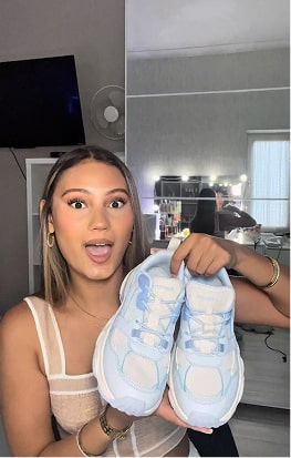 Person holding a pair of light blue sneakers, smiling in a room with a mirror.