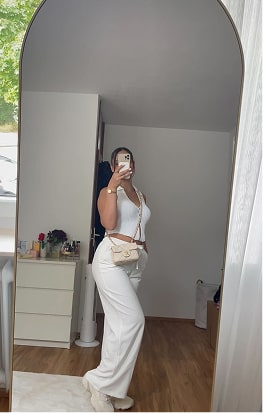Person in white outfit taking a mirror selfie in a room with wooden floor.