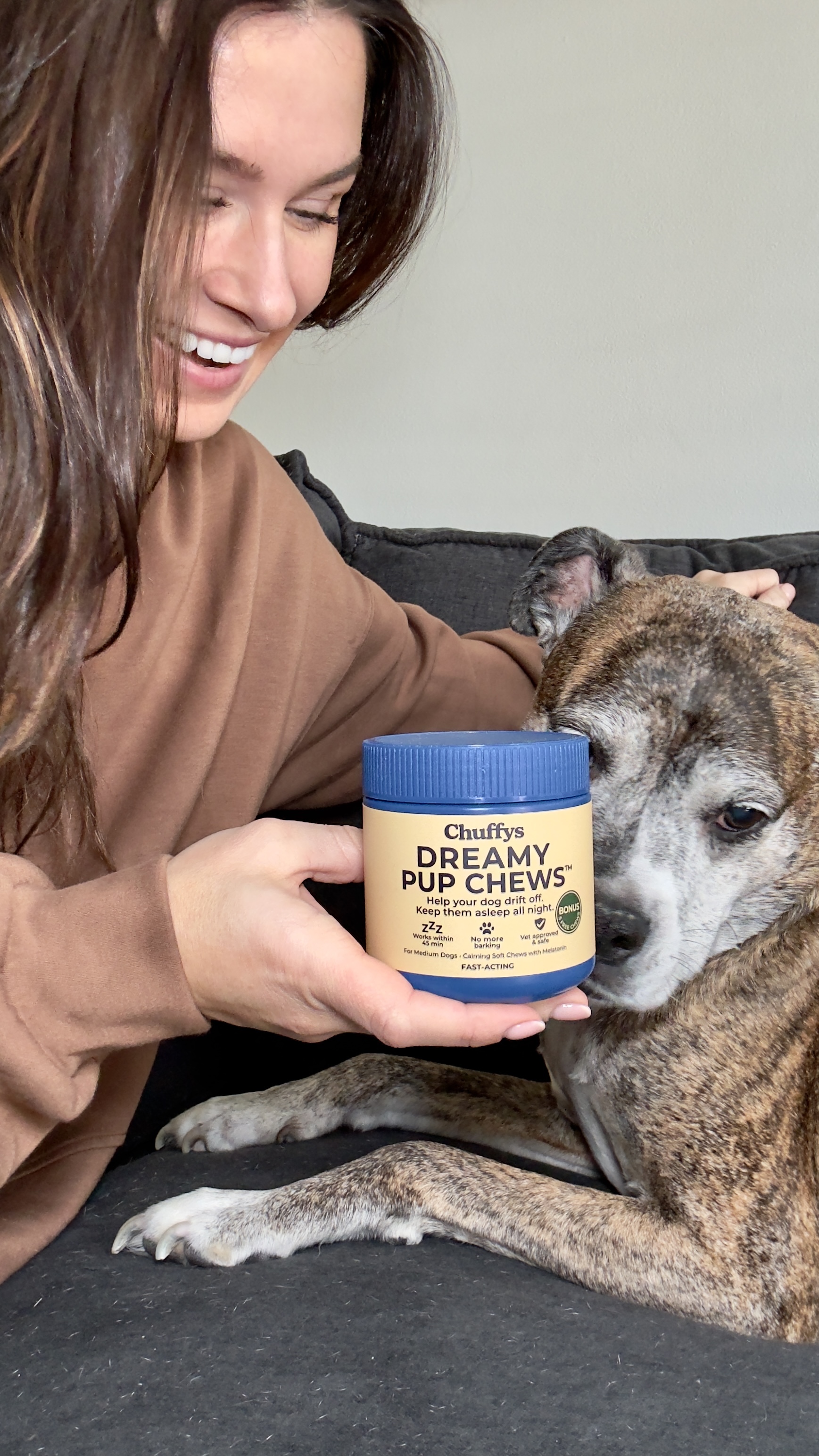 A smiling woman holds a jar of Chuffys Dreamy Pup Chews next to a brindle dog she's petting.