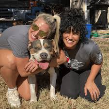 Two smiling women kneel on the grass and hug a happy-looking brown and white dog between them.