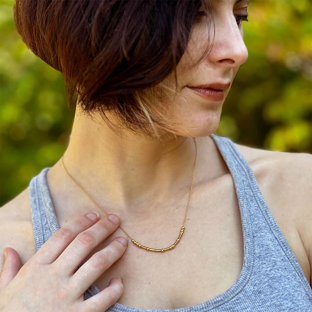 A woman with short brown hair wearing a grey tank top and a gold morse code style necklace.