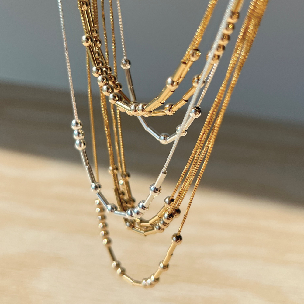 A close-up of several layered gold and silver necklaces with a beaded bar design.