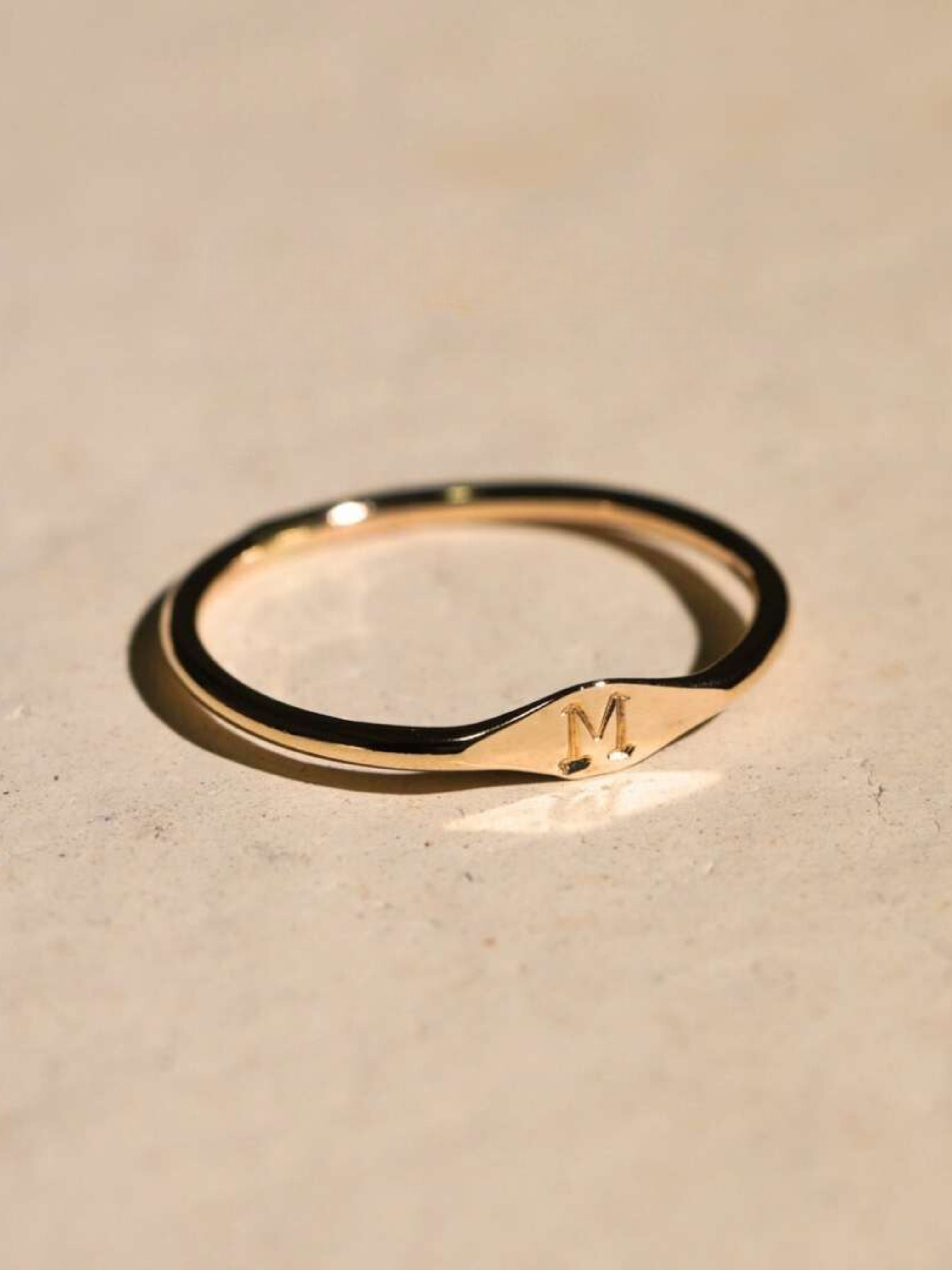 A delicate gold signet ring with the initial 'M' engraved on it, resting on a light surface.