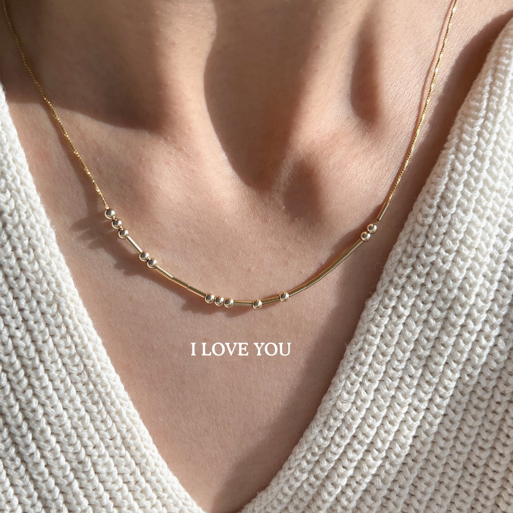 A close-up of a gold necklace with beads and bars spelling 'I LOVE YOU' in Morse code.
