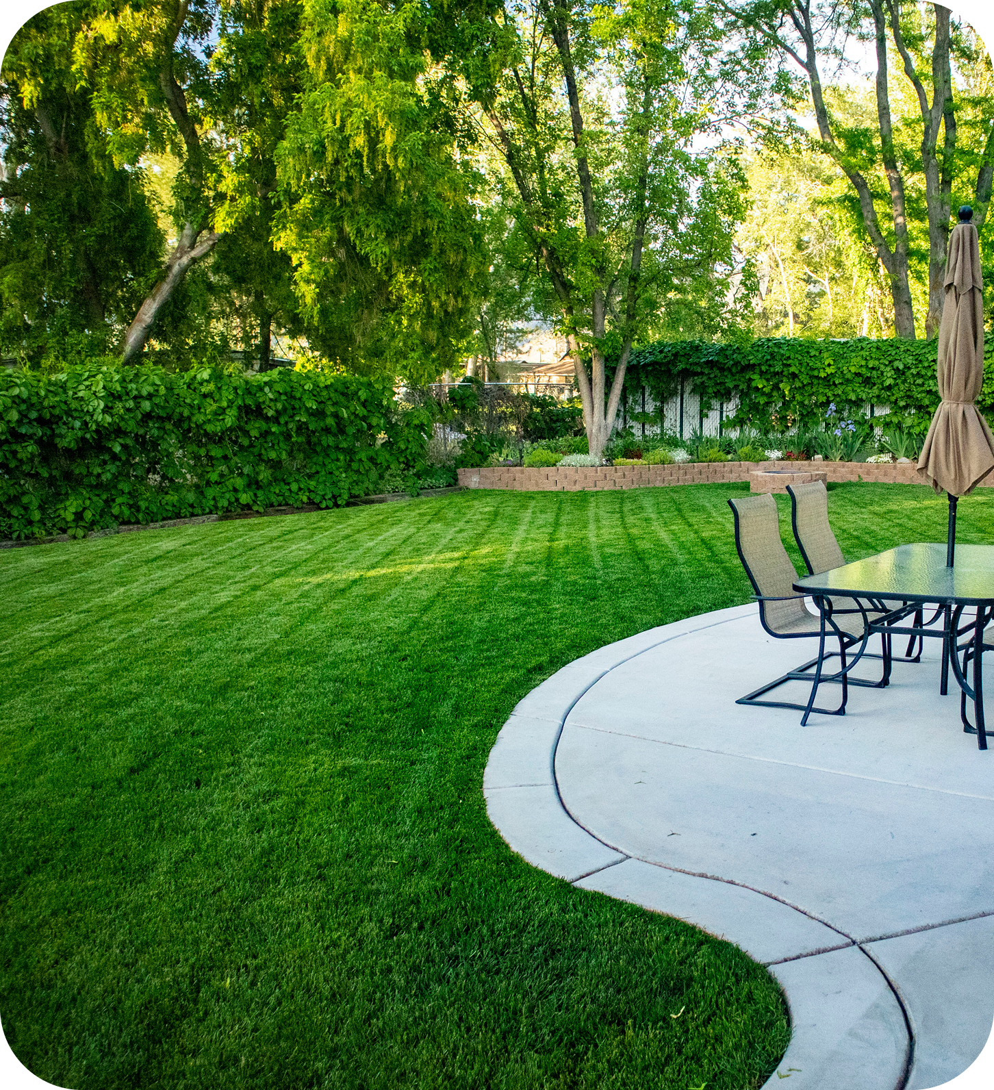 A backyard with a patio, table, chairs, and lush green lawn surrounded by trees.