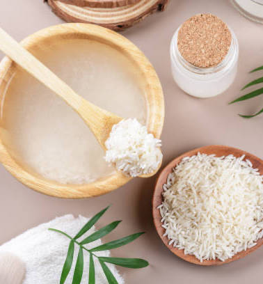 An overhead shot of wooden bowls with raw and cooked rice, a jar, and a palm frond.