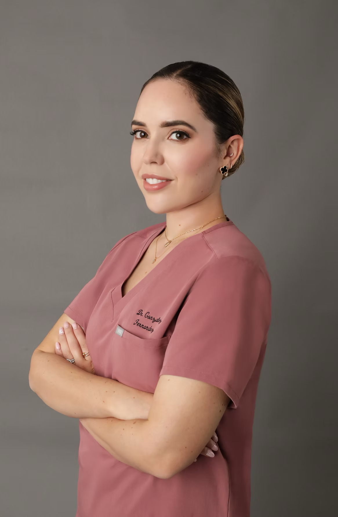 Person in pink medical scrubs, arms crossed, smiling against a gray background.