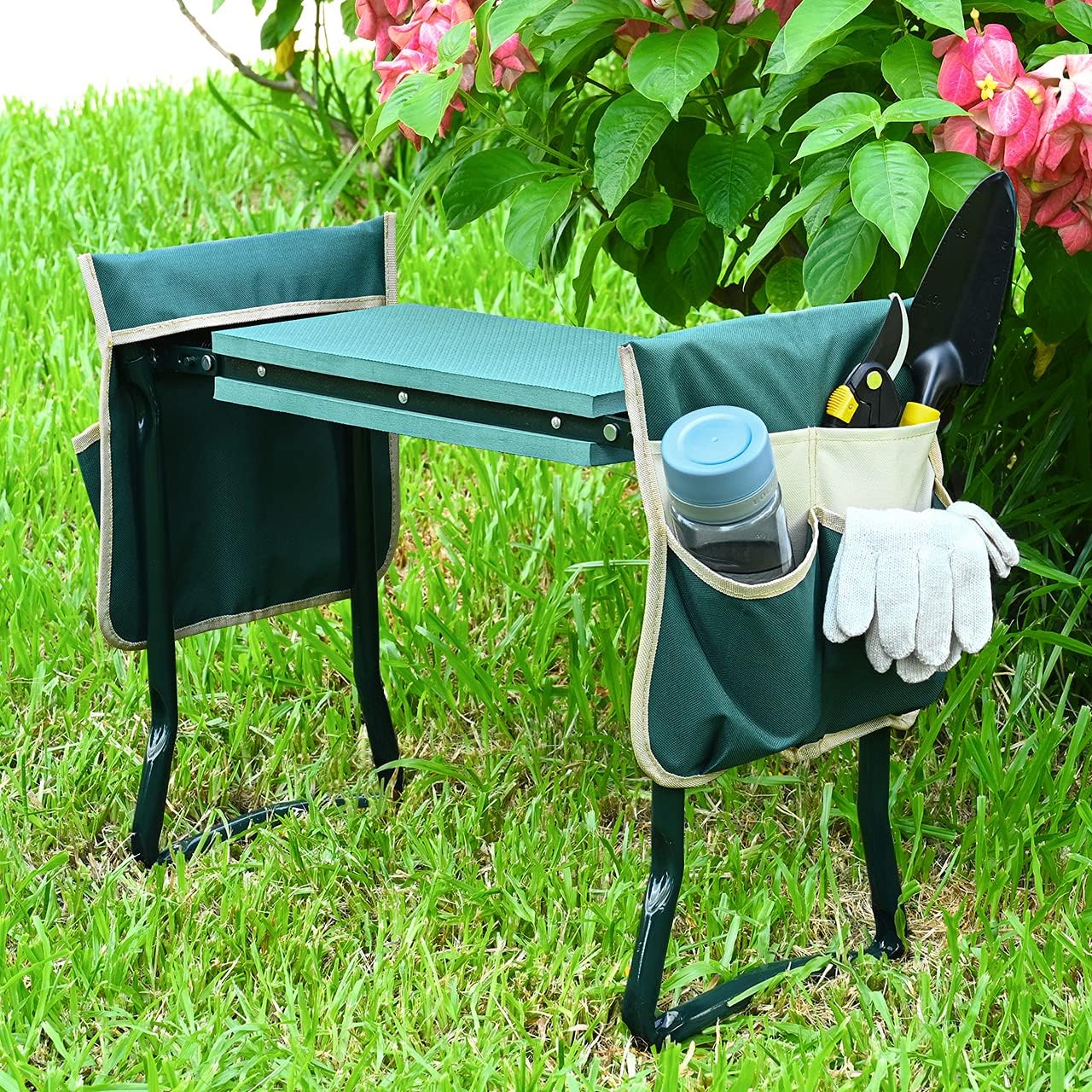 A green gardening kneeler and seat with tool pouches sits on a lawn next to pink flowers.