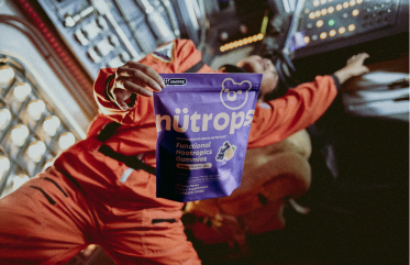 A person in an orange jumpsuit holds up a purple pouch of nütrops gummies inside a spaceship.