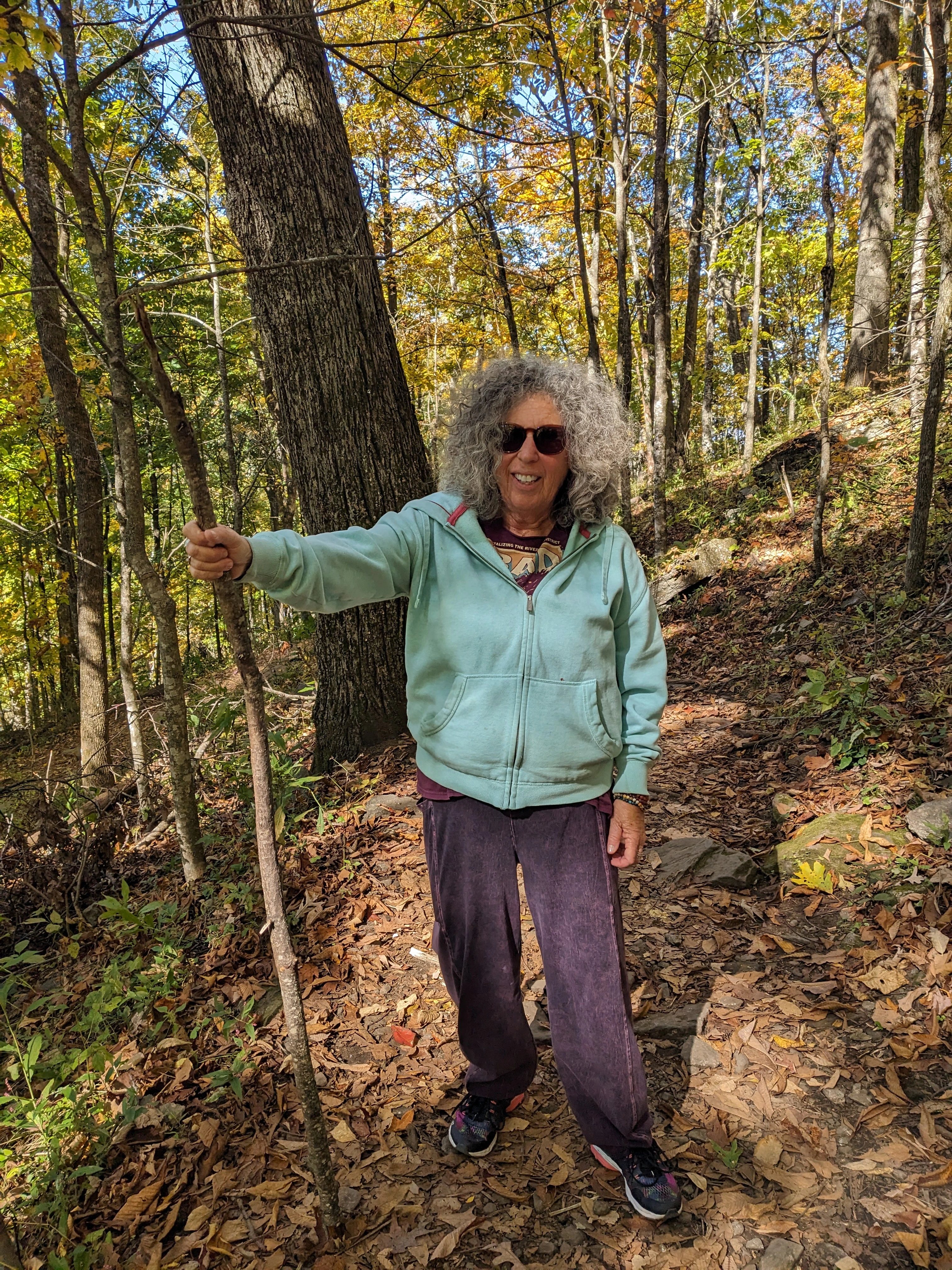 A woman with curly gray hair and sunglasses smiles while hiking on a wooded, leaf-covered trail.