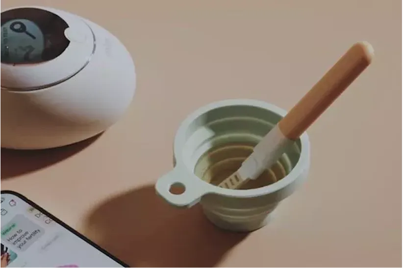 A hormone test stick in a collapsible cup, next to a digital reader and a smartphone.