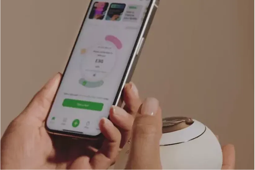 A person holds a smartphone displaying an app next to a small, round, white electronic device.