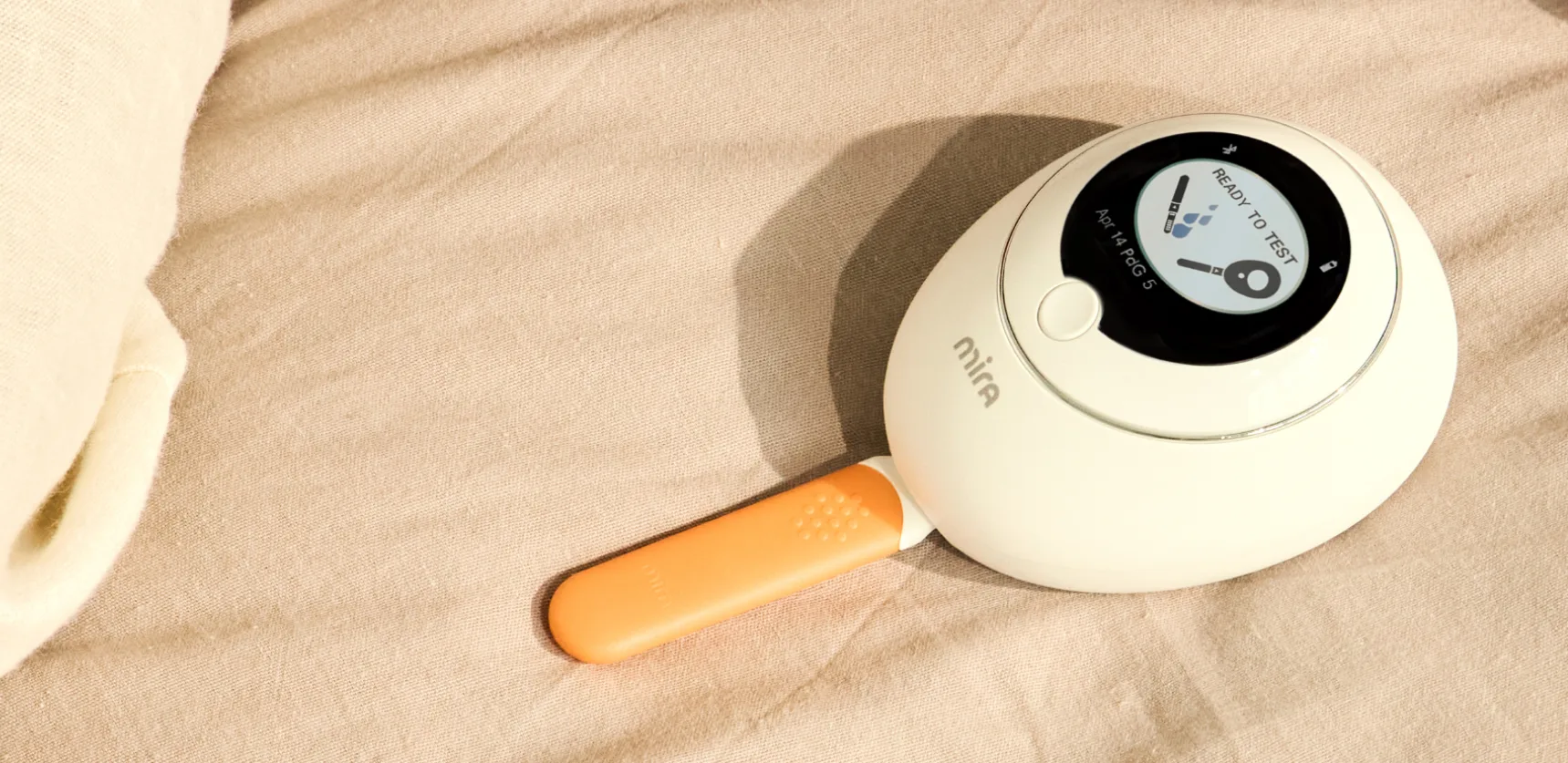 A white Mira digital fertility analyzer with an orange test wand inserted, resting on a beige fabric.
