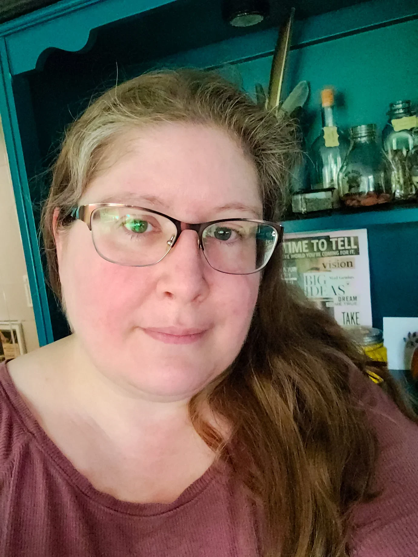Person with glasses, long hair, in front of a shelf with bottles and decor.