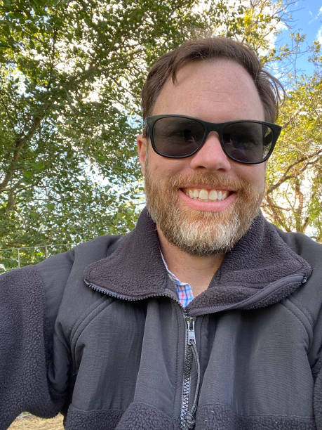 Person smiling, wearing sunglasses and a jacket, in an outdoor setting with trees in the background.