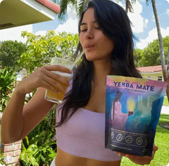 Woman drinking a beverage, holding a bag of yerba mate outside in a garden.
