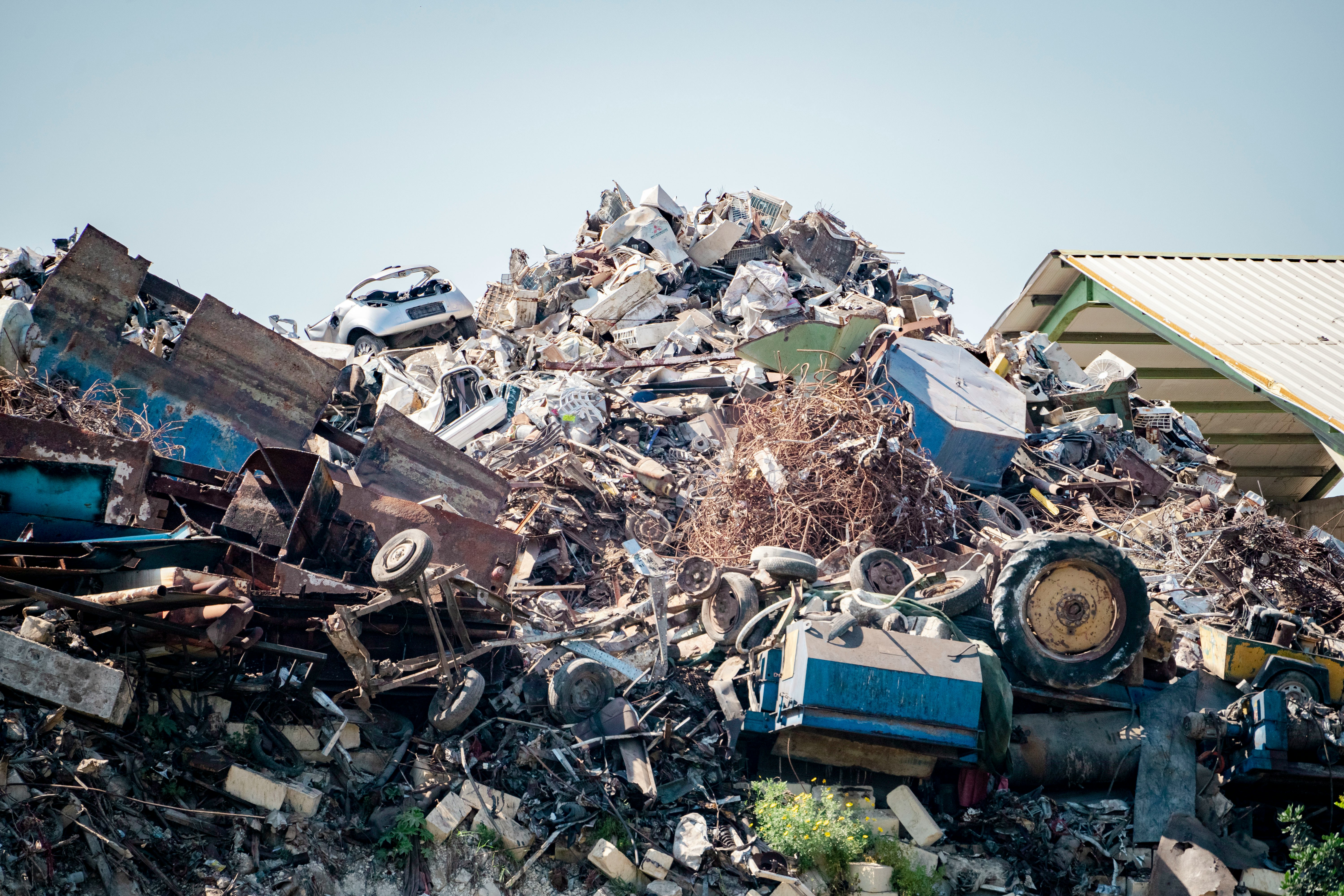 A large pile of scrap metal, including wrecked cars and tires, sits in a junkyard under a clear sky.