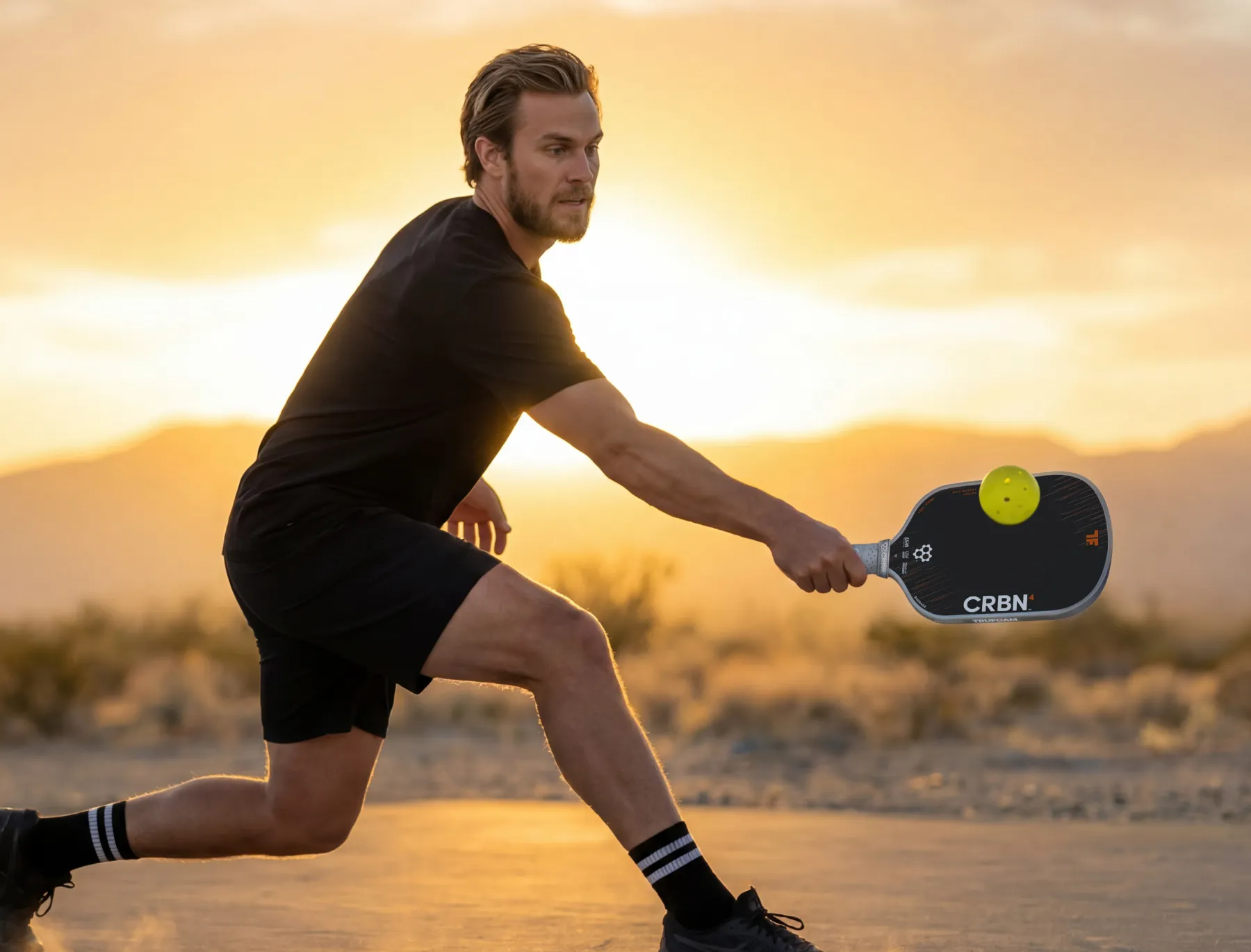 A man in black sportswear lunges to hit a yellow pickleball with a paddle outdoors at sunset.