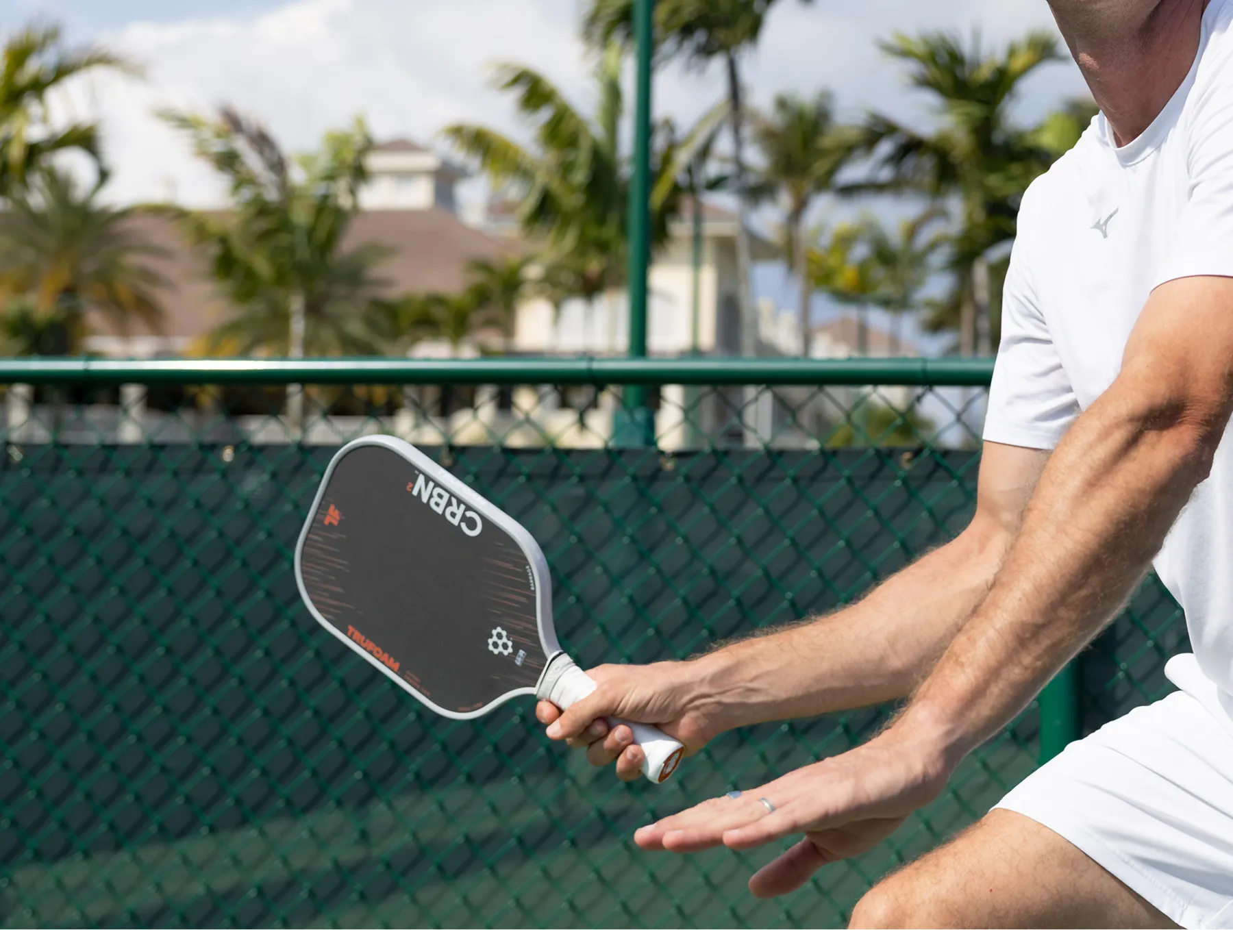 A man in white sportswear holds a black pickleball paddle on an outdoor court with palm trees.