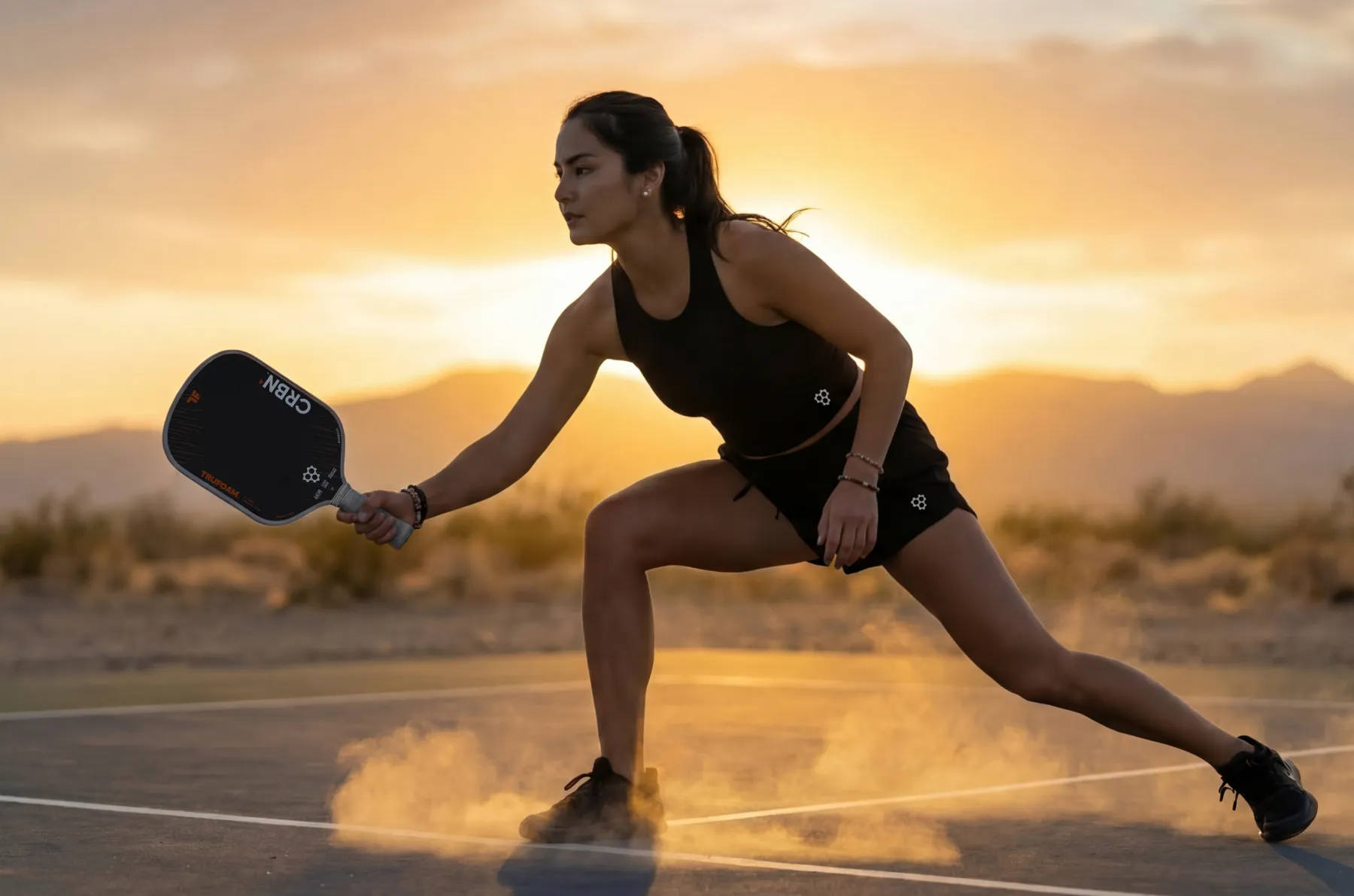 A woman lunges on a pickleball court with a paddle, kicking up dust at sunset.