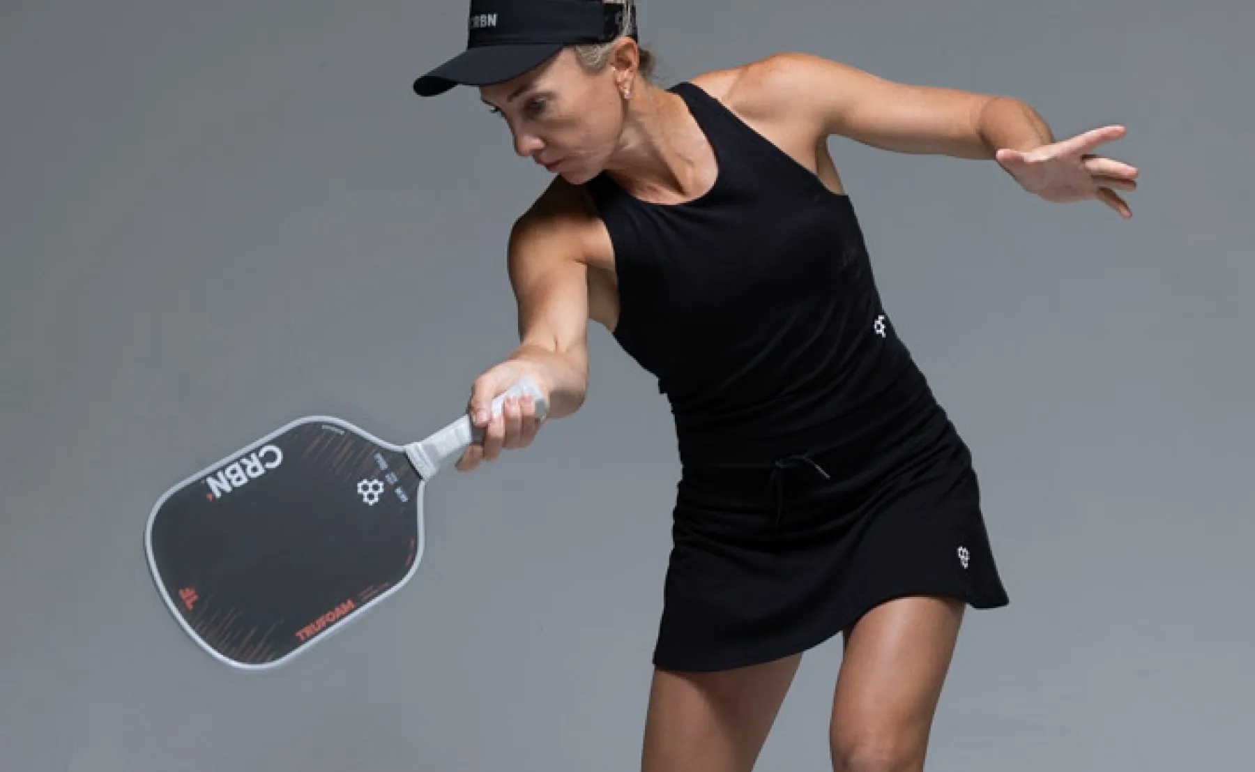 A woman in black athletic wear and a visor swings a CRBN pickleball paddle against a grey background.