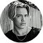 A black and white circular headshot of a young man with medium-length blonde hair and a chain necklace.