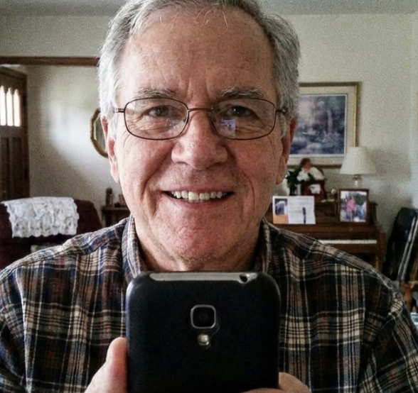 An older man with glasses and a plaid shirt takes a selfie with his phone in a living room.