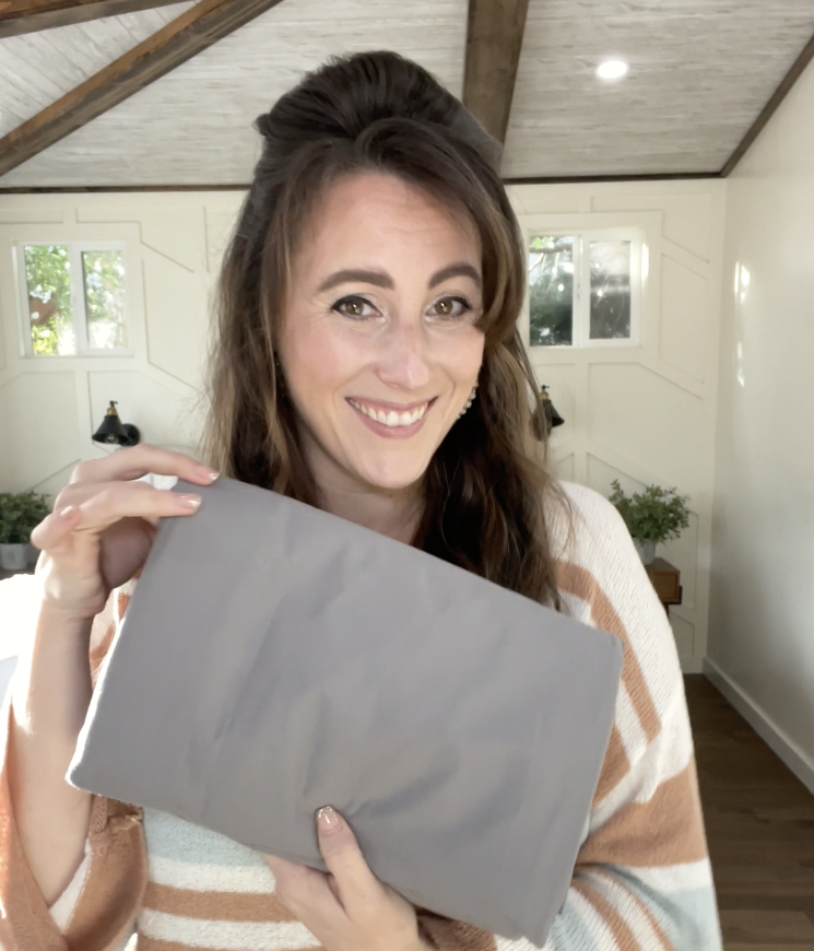 Person smiling and holding a folded gray fabric inside a room with beams and bright windows.
