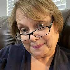 Person wearing glasses, sitting on a brown leather couch with blinds in the background.