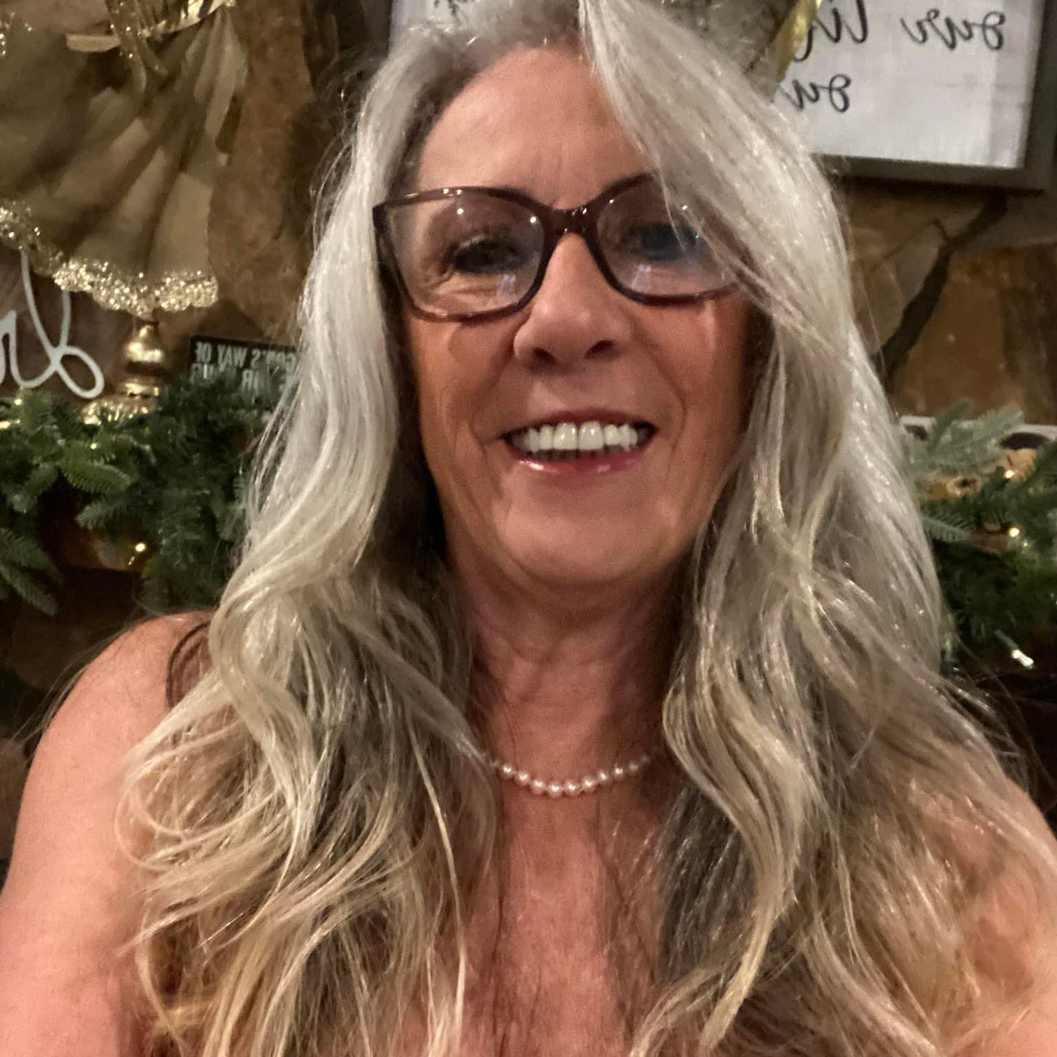 A smiling woman with long, wavy gray hair, glasses, and a pearl necklace takes a selfie.