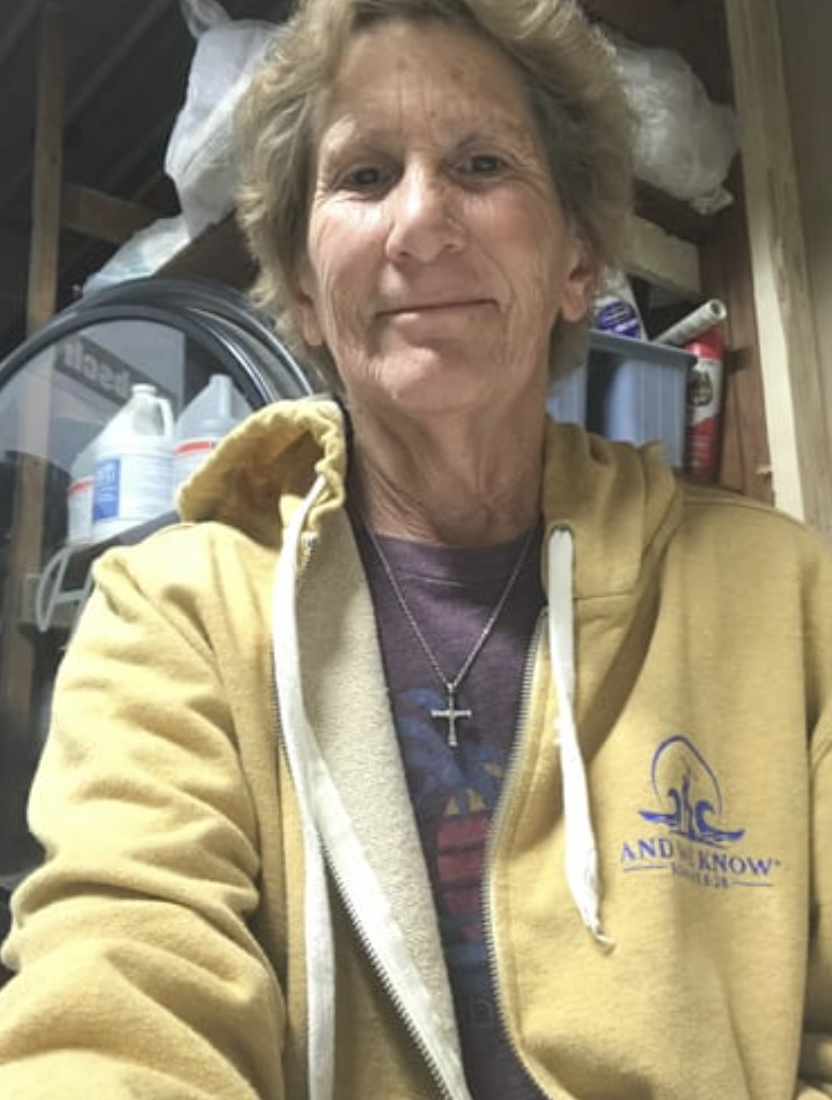 Person in a yellow jacket with a cross necklace, standing indoors with shelves in the background.