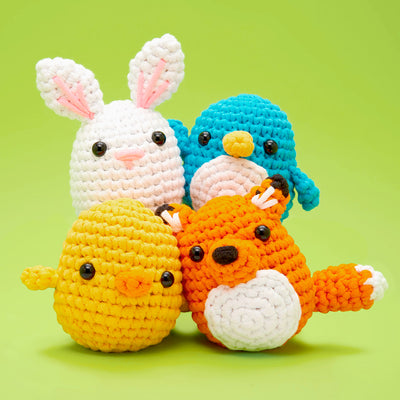 Four colorful crocheted stuffed animals, a bunny, chick, fox, and penguin, grouped on a green background.