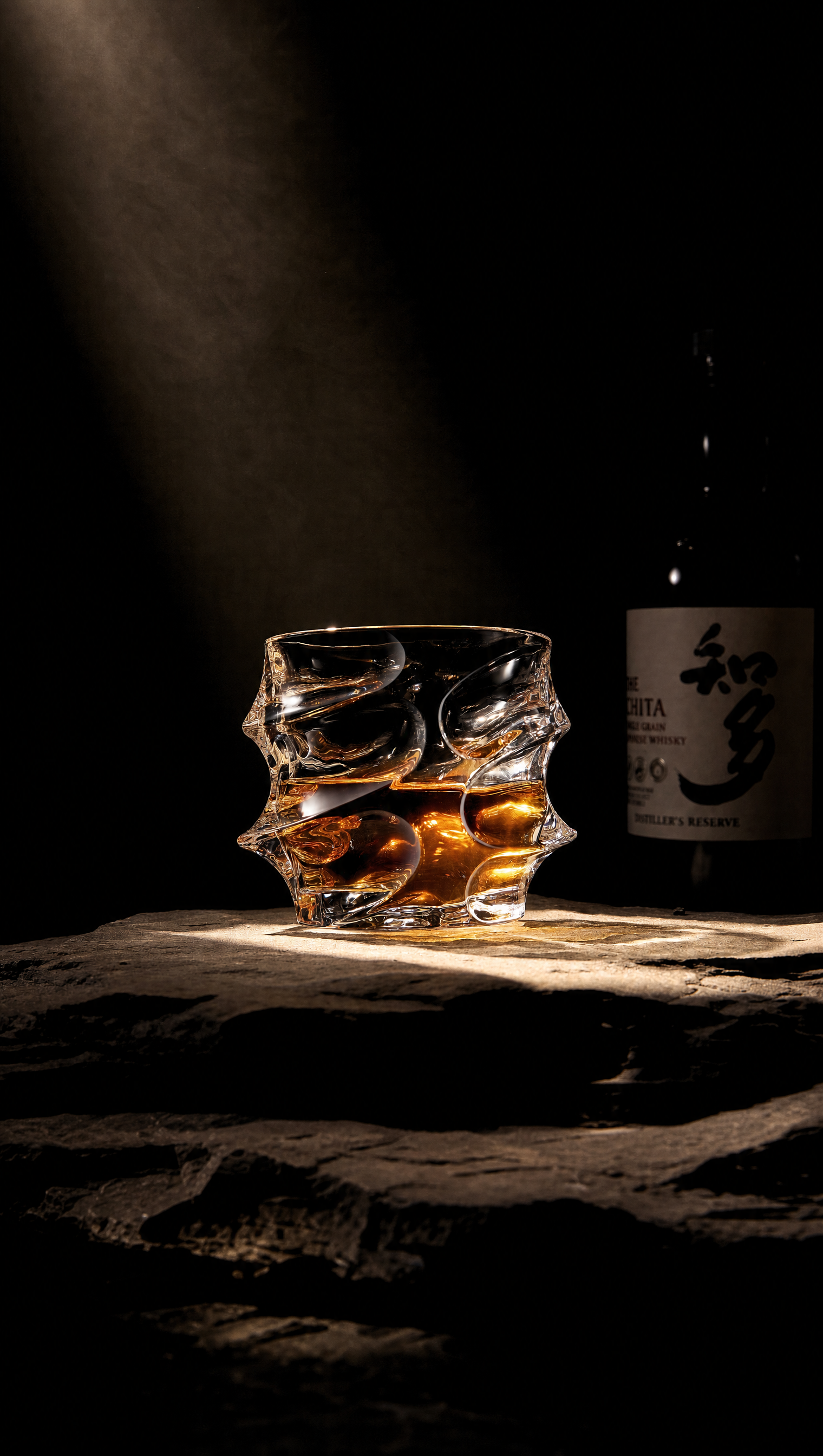 A uniquely shaped glass of whisky sits on a stone surface, dramatically lit, with a bottle behind it.