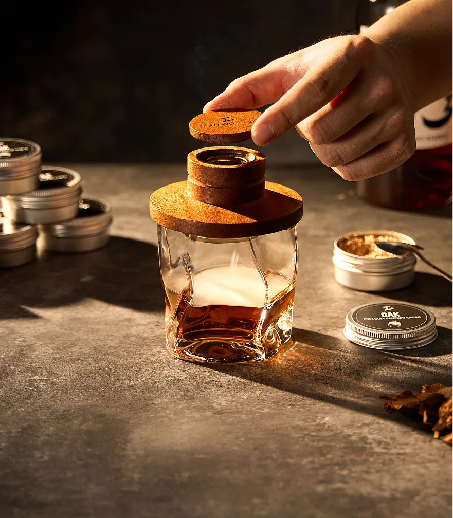 A hand places a wooden smoker lid on a glass of liquor next to tins of wood chips.