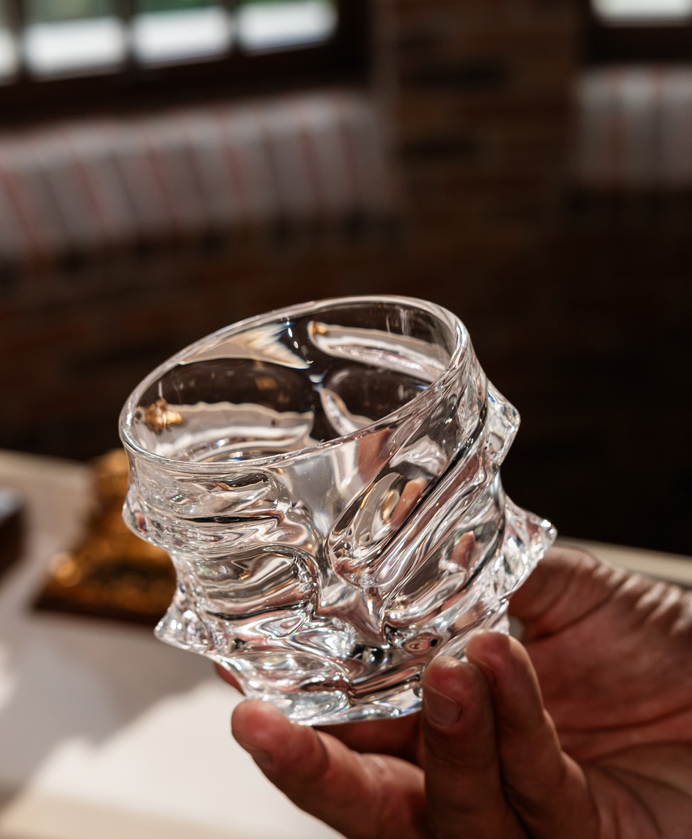 A hand holding an abstract, textured glass cup indoors.