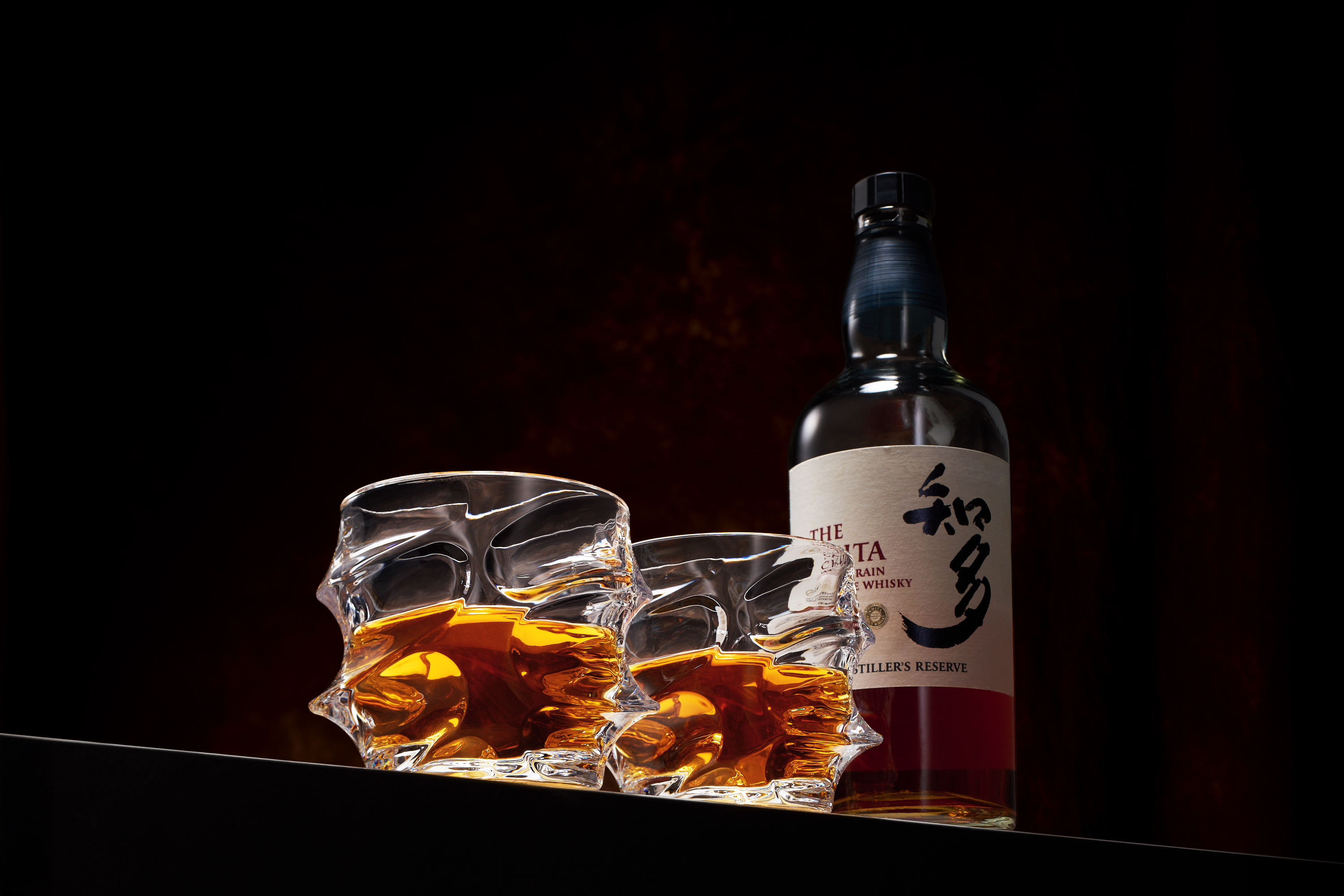 A bottle of Japanese whisky and two uniquely shaped glasses filled with the liquid against a dark background.