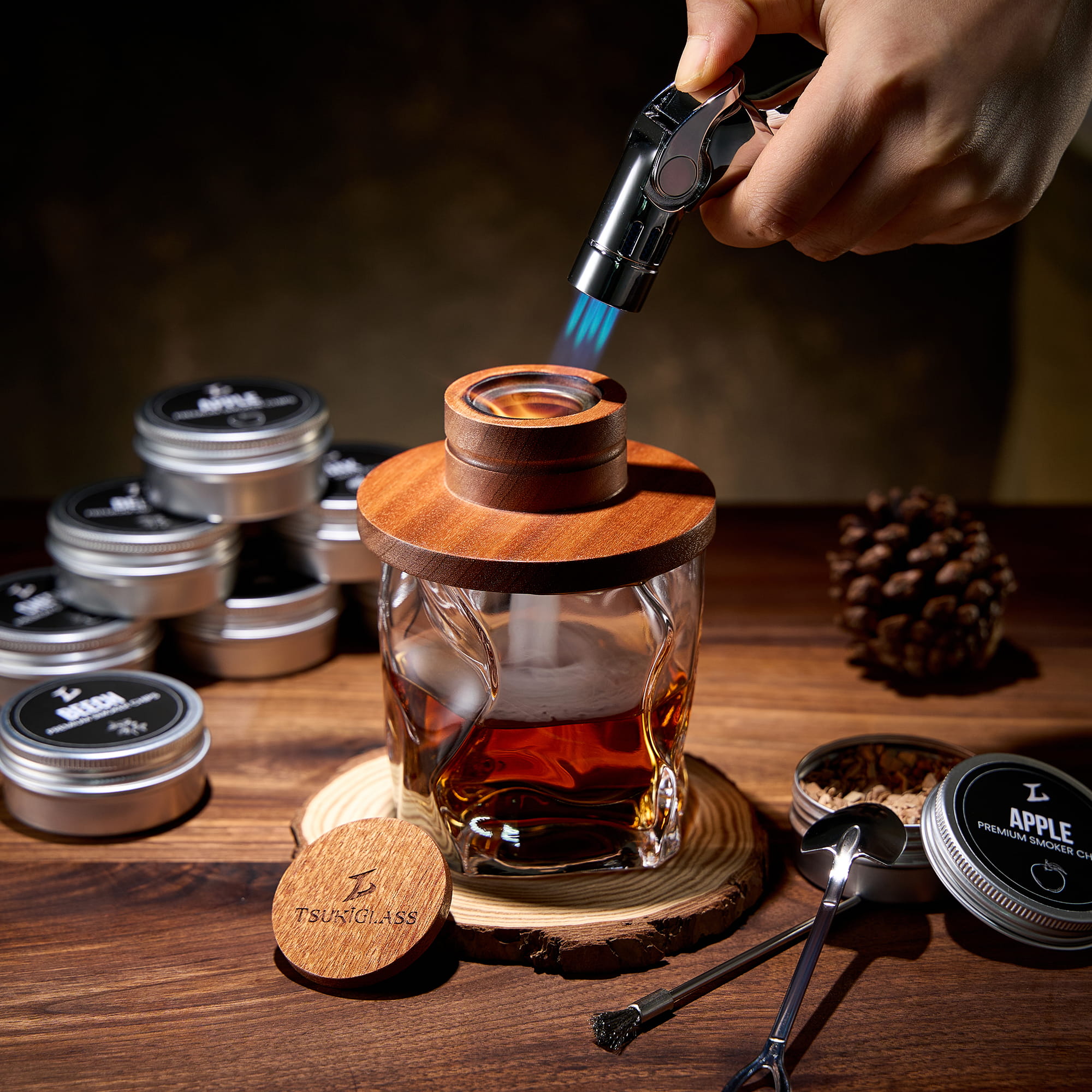 A hand holds a torch over a cocktail smoker on a glass of whiskey on a wooden table.