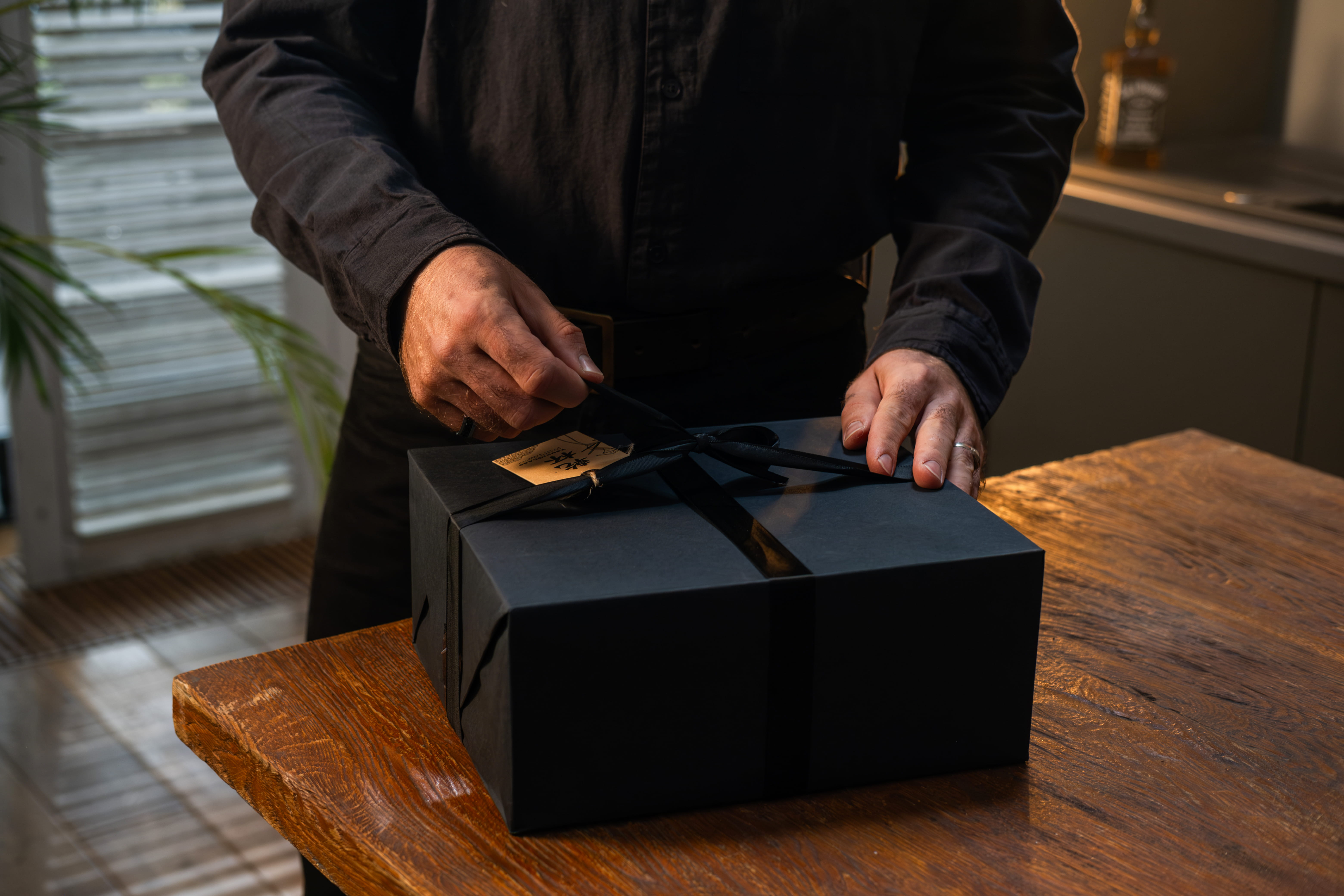 A person in a black shirt ties a ribbon on a black gift box on a wooden table.