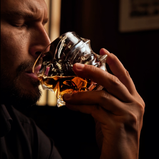 A man sips an amber liquid from an ornate, uniquely shaped glass in a dimly lit setting.